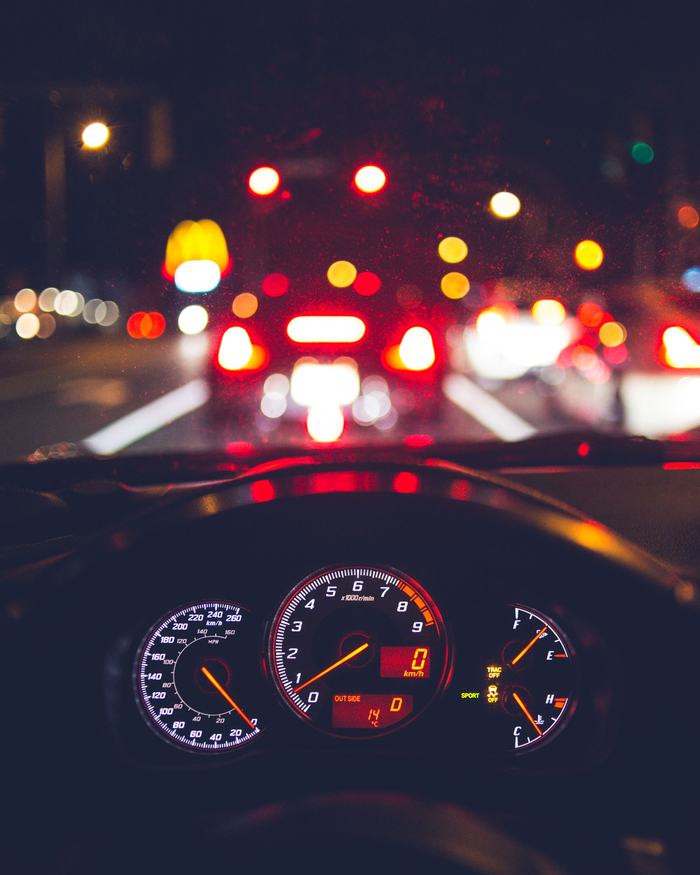 A smartphone showing a map app on a car dashboard at night, with city traffic blurred beyond the windshield